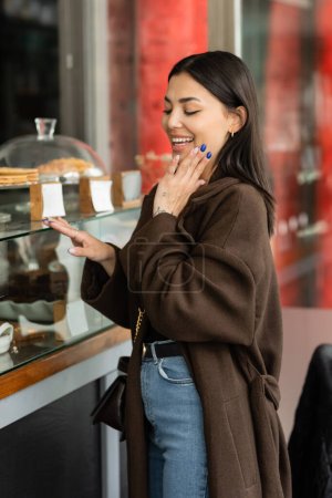 mujer asombrada sosteniendo la mano cerca de la cara mientras mira escaparate de pastelería con postres en praga 