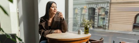 brunette woman in coat drinking coffee and looking through window in prague cafe, banner