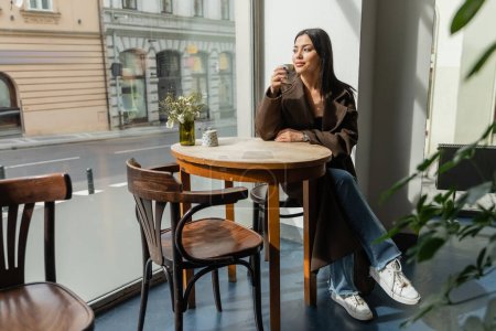 full length of brunette woman looking at city street while sitting in cafe in prague