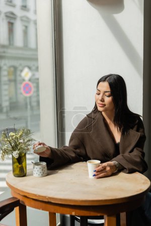 brünette Frau mit Pappbecher öffnet Zuckerdose, während sie in der Nähe des Fensters im Prager Café sitzt