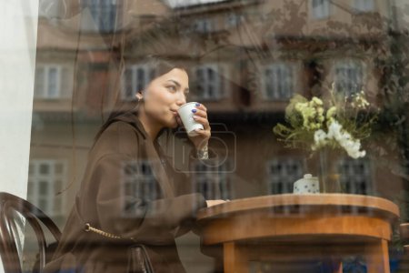 junge Frau im Herbstmantel trinkt Kaffee am Fenster eines Prager Cafés