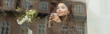 Lächelnde Frau mit Einwegbecher blickt durch Fenster in Prager Café, Banner