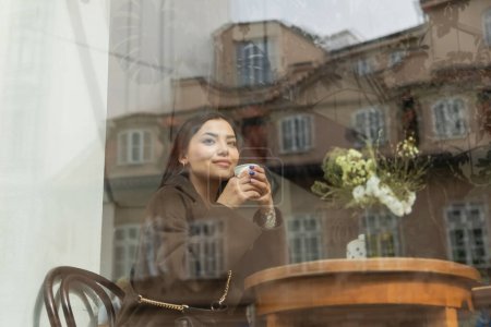 junge brünette Frau im Mantel trinkt Kaffee aus Pappbecher in der Nähe des Fensters im Prager Café