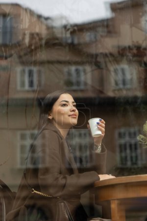 Brünette Frau im Mantel hält Pappbecher in der Hand und schaut am Fenster in Prager Café weg