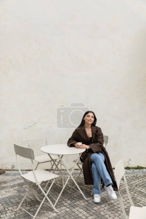 Smiling woman in jeans and coat holding paper cup in outdoor cafe in Prague 