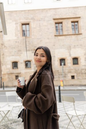 Téléchargez les photos : Portrait de jeune femme en manteau tenant une tasse en papier et souriant à la caméra dans la rue à Prague - en image libre de droit