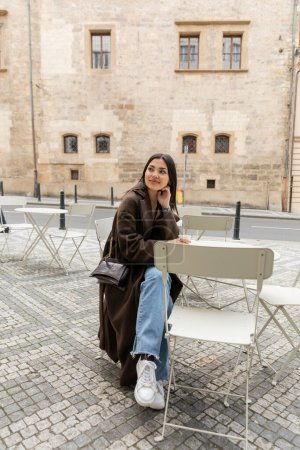 Trendige junge Frau im Mantel schaut in der Nähe einer Papptasse mit Kaffee in einem Café in Prag weg 