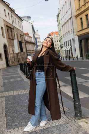 Joyful woman in coat holding paper cup on urban street in Prague 