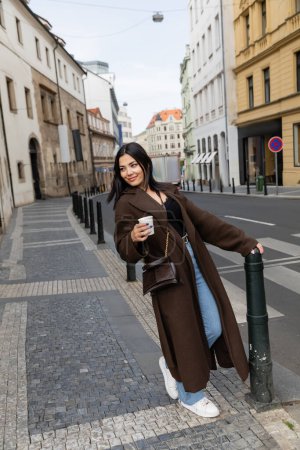 Happy tourist in coat holding paper cup on street in Prague 