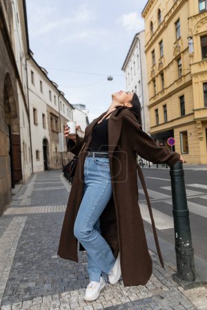 Side view of joyful woman in coat holding paper cup on street in Prague 