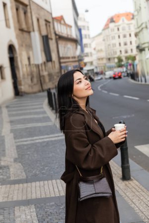 Young brunette woman in coat with handbag holding paper cup near road on urban street in Prague 