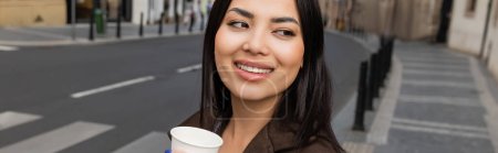 Smiling brunette woman in coat holding paper cup and looking away on blurred urban street, banner 