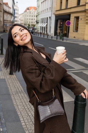 Positiver Reisender im Mantel hält Pappbecher in der Nähe der Straße auf verschwommener Stadtstraße in Prag 