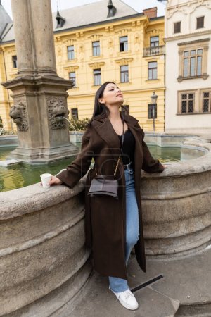 Elegante turista en abrigo sosteniendo taza de papel cerca de la fuente en la Plaza Charles en Praga 