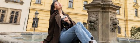 Téléchargez les photos : Femme agréable en manteau tenant une tasse en papier près de la fontaine sur la place Charles à Prague, bannière - en image libre de droit