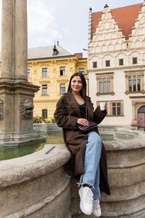 Happy traveler in coat holding paper cup near ancient fountain in Charles Square in Prague 