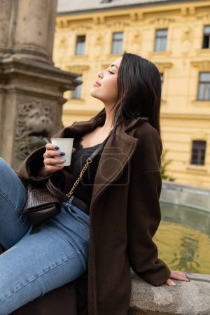 Téléchargez les photos : Vue latérale d'une femme élégante en manteau tenant une tasse en papier près d'une fontaine sur la place Charles à Prague - en image libre de droit