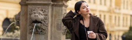 Téléchargez les photos : Élégante jeune femme en manteau tenant une tasse en papier près d'une fontaine floue sur la place Charles à Prague, bannière - en image libre de droit