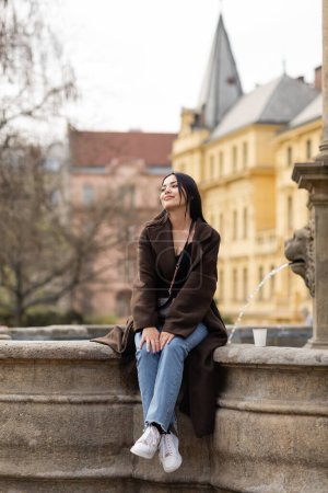 Foto de Mujer sonriente con abrigo marrón mirando hacia otro lado cerca de la taza de papel en la Plaza Charles en Praga - Imagen libre de derechos