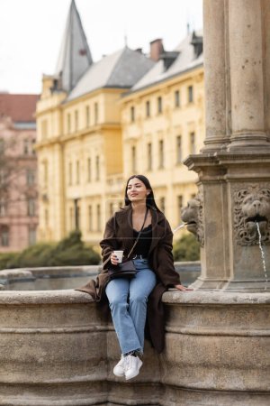 Smiling woman in coat holding paper cup while sitting on ancient fountain in Charles Square in Prague 