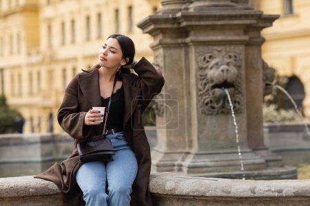Junge Frau im Mantel berührt Haare und hält Pappbecher in der Nähe des verschwommenen Brunnens auf dem Prager Karlsplatz 