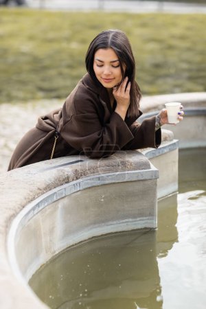 Brunette woman in coat holding paper cup near fountain in Prague