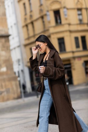Seitenansicht eines stilvollen Touristen in Mantel und Sonnenbrille mit Kaffee zum Mitnehmen auf der Straße in Prag 