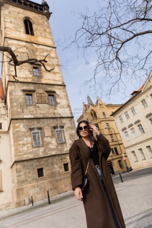 Téléchargez les photos : Femme élégante en manteau et lunettes de soleil debout près des bâtiments à Prague - en image libre de droit