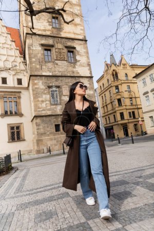 Stilvolle Frau in Sonnenbrille und Mantel läuft auf der Straße in Prag 