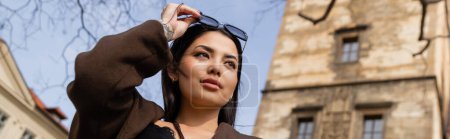 Low angle view of stylish brunette woman holding sunglasses on street in Prague, banner 