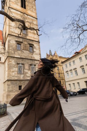 Téléchargez les photos : Vue latérale de la femme souriante en manteau tenant des lunettes de soleil tout en tournant dans la rue à Prague - en image libre de droit