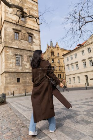 Young woman in coat holding sunglasses while walking on urban street in Prague 