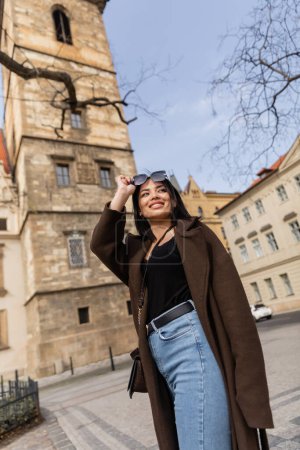 Low angle view of happy traveler in coat holding sunglasses near blurred buildings in Prague 