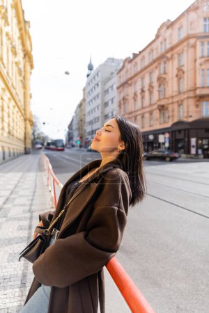 Side view of brunette woman in coat standing near railing on street in Prague 