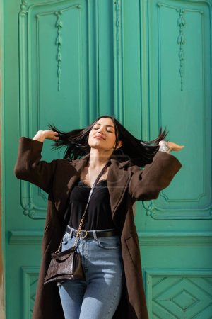 Téléchargez les photos : Jeune femme en manteau touchant les cheveux près de la porte turquoise du bâtiment sur la rue urbaine - en image libre de droit