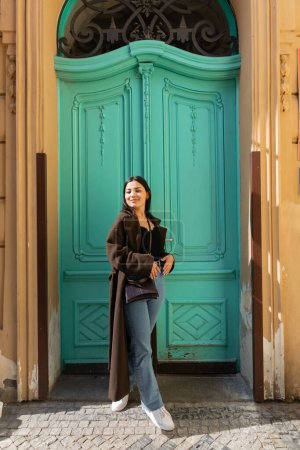 Lächelnde Frau im Mantel blickt in die Kamera in der Nähe der türkisfarbenen Tür eines Gebäudes auf der Straße in Prag 
