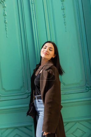 Téléchargez les photos : Femme agréable en manteau debout dans la lumière du soleil près de la porte turquoise de l'immeuble sur la rue - en image libre de droit