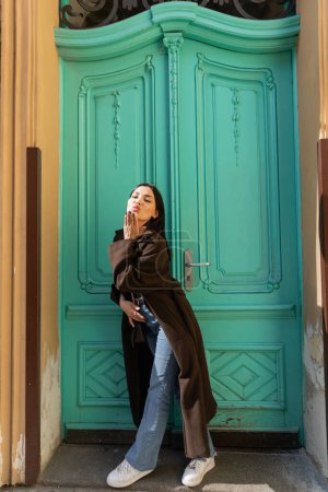 Woman in coat blowing air kiss at camera near turquoise door of building on street 