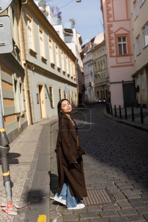 Brunette woman in coat walking on blurred urban street in Prague 
