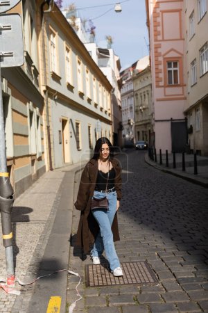 Smiling traveler in brown coat walking on road on urban street in Prague 