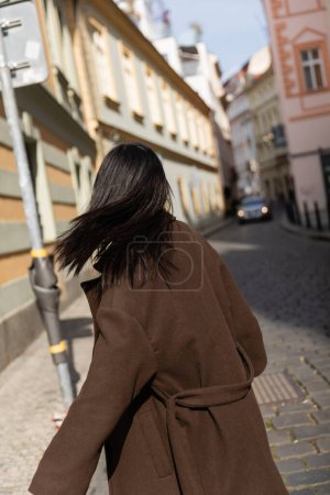 Back view of brunette woman in coat standing in blurred street in Prague 