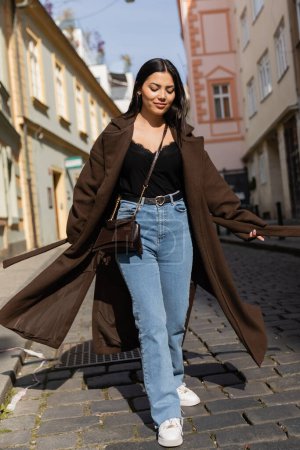 Mujer sonriente en abrigo caminando por carretera en la calle urbana de Praga 