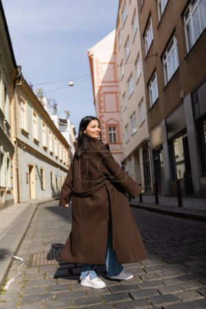 Téléchargez les photos : Femme positive en manteau debout dans la lumière du soleil sur la rue à Prague - en image libre de droit