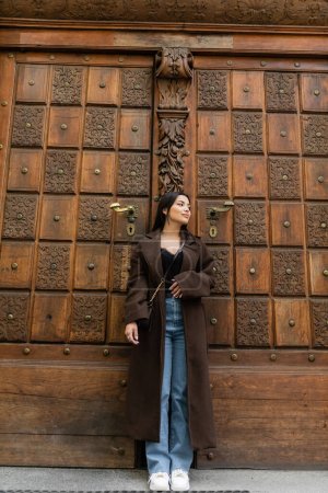 Stylish woman in coat standing near antique door of building on street in Prague 