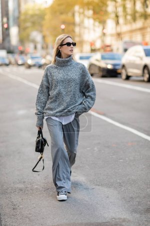 Téléchargez les photos : Pleine longueur de femme blonde en lunettes de soleil et tenue grise tenant sac à main tout en marchant sur la rue de New York - en image libre de droit