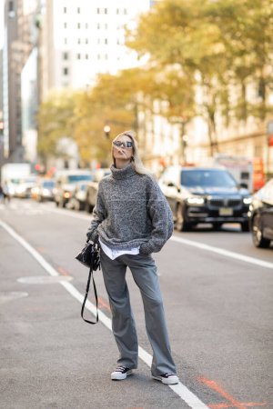 longitud completa de la mujer con estilo en gafas de sol y traje gris sosteniendo bolso negro y de pie en la calle de la ciudad de Nueva York 