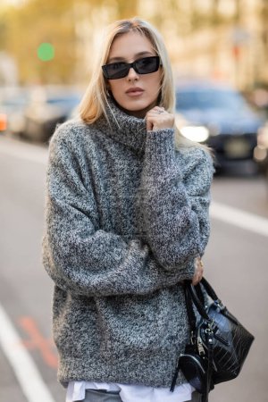Schöne Frau in Sonnenbrille und grauem Outfit mit Handtasche in New York City 