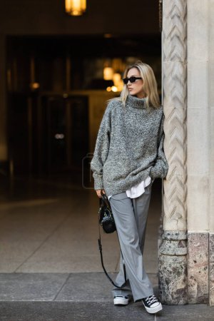 Téléchargez les photos : Pleine longueur de femme blonde en lunettes de soleil et tenue grise tenant le sac à main tout en se tenant près du bâtiment à New York - en image libre de droit