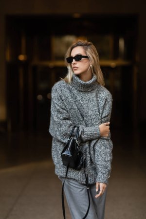 mujer con estilo en gafas de sol y suéter gris de pie con bolso negro en Nueva York 