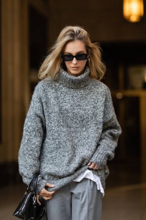 stylish woman in sunglasses and grey sweater walking with black handbag in New York 
